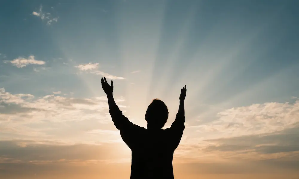A person with hands slightly raised in quiet worship, silhouette against an expansive sunrise sky.