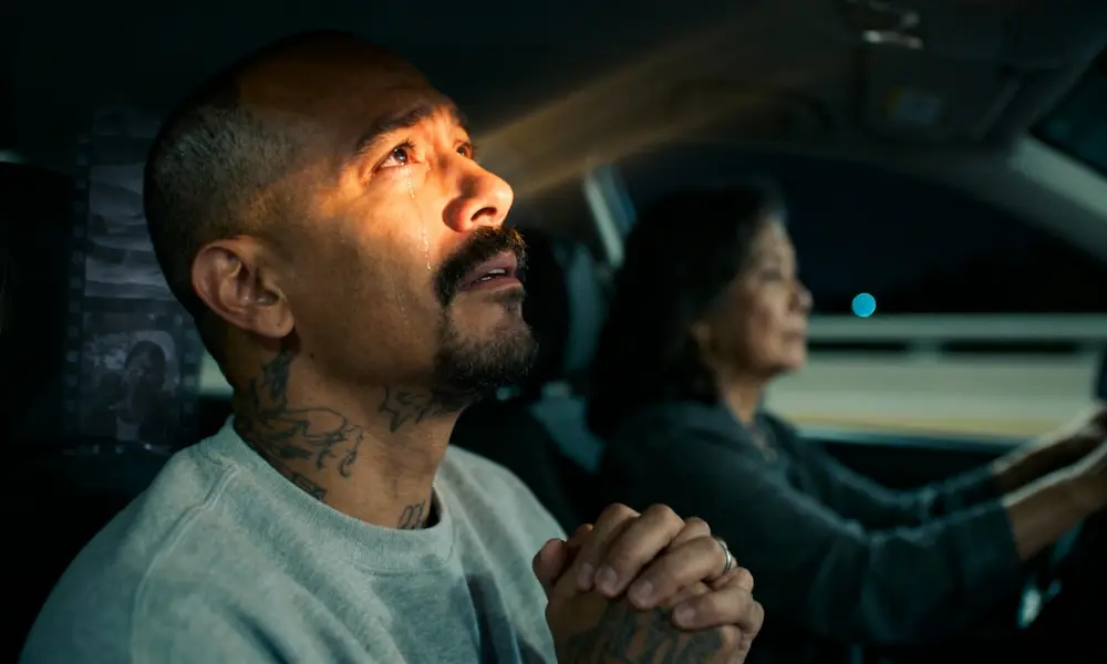 Middle-aged man in a car at night breaking down as a warm, ethereal light touches his face, faint flashback of layered darkness behind him, mother driving.