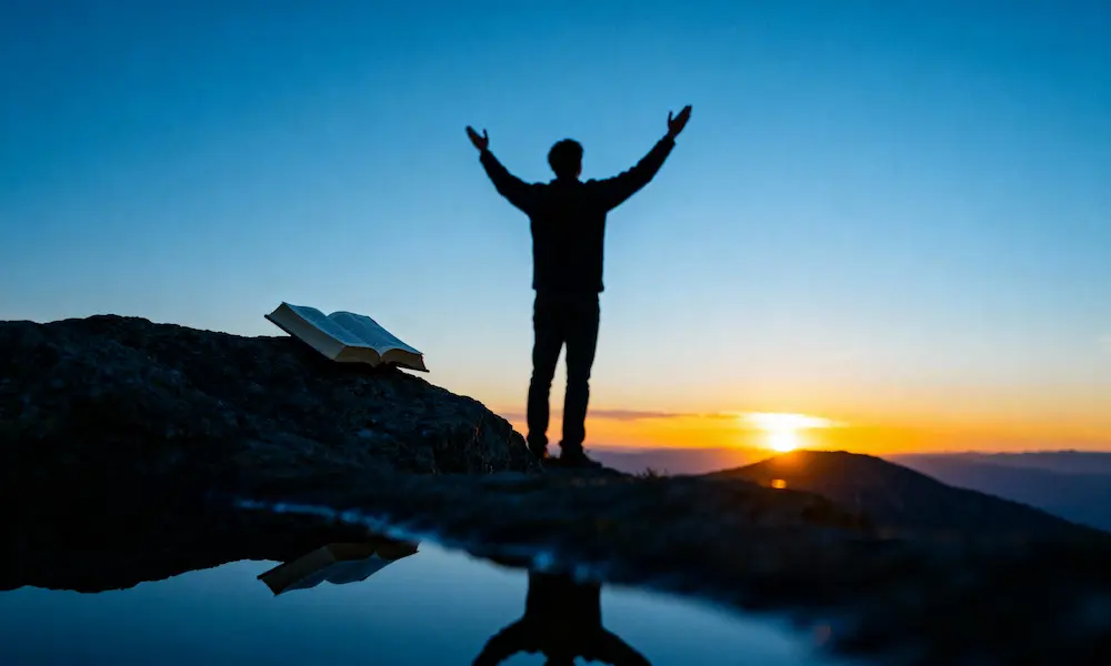 Person stands with raised arms at mountain overlook at blue hour, Bible on nearby rock.