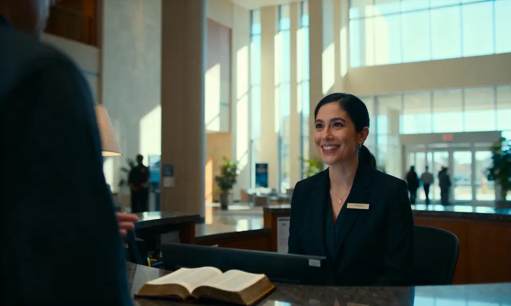 Professional receptionist greets visitors warmly with open Bible on desk and morning lobby light.