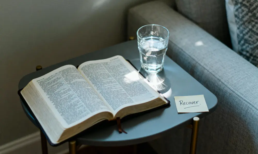 Open Bible on side table with water and “Recover” note in soft daylight.