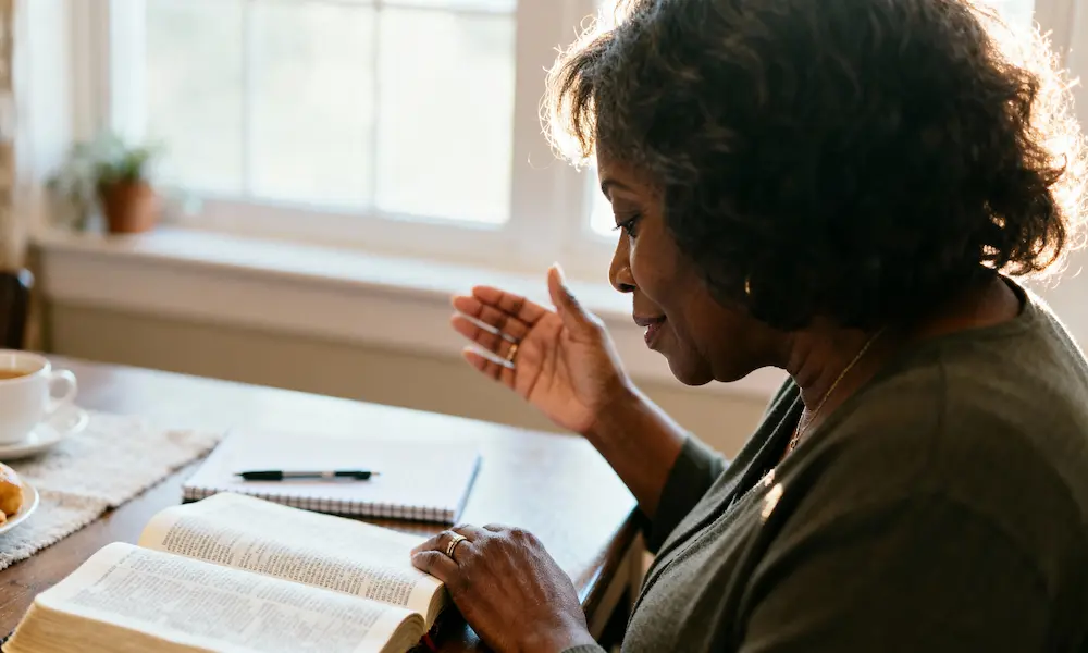 Grandmother at breakfast nook with open Bible, hand lifted in thanks.