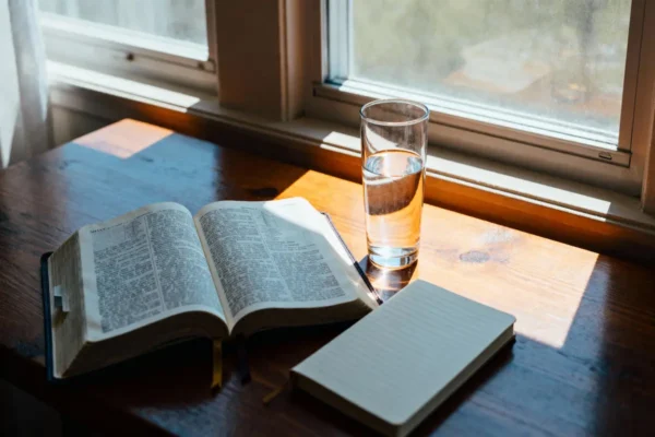 Open Bible by a bright window with journal and water, warm light and ample copy space.