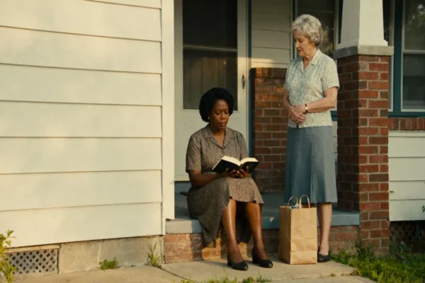 Neighbors on a stoop with an open Bible and groceries, late-morning sun.
