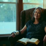 Woman with wrist brace by window, open Bible on lap, warm light and copy space.