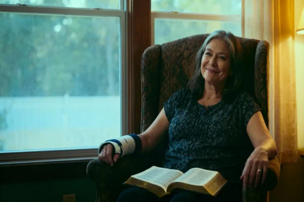 Woman with wrist brace by window, open Bible on lap, warm light and copy space.